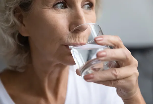 Ältere Frau trinkt aus einem Glas Wasser, Seitenansicht, Hände halten das transparente Trinkglas vor dem Mund, Fokus auf Gesicht und Hände.