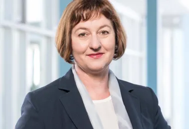 Porträt einer Frau mit schulterlangen, braunen Haaren, die einen dunkelblauen Blazer und einen hellen Schal trägt. Sie steht in einem hellen Raum mit großen Fenstern im Hintergrund und lächelt leicht.