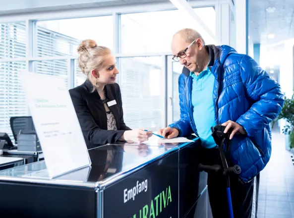 Empfangsbereich eines Ambulatoriums mit einer jungen Frau in schwarzer Bluse, die einem älteren Mann in blauer Jacke Informationen gibt. Der Mann hält einen Gehstock und schaut auf ein Formular. Im Hintergrund sind moderne Büromöbel und große Fenster s...