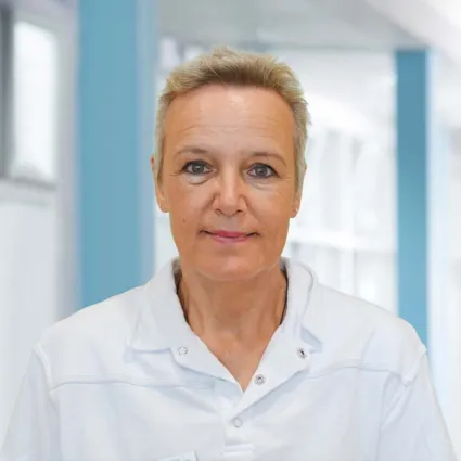 Frau mit kurzen, blonden Haaren und einem weißen Oberteil, lächelt in einem hellen, modernen Raum mit blauen Akzenten.