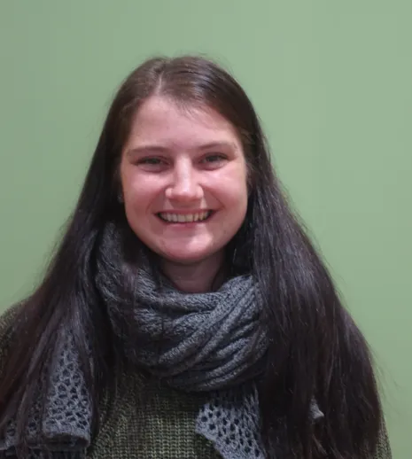 Lächelnde Frau mit langen, dunklen Haaren, die einen grauen Schal und einen grünen Pullover trägt, vor einer hellgrünen Wand.