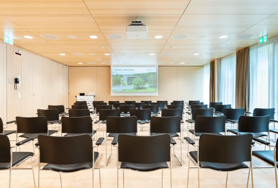 Leerer Konferenzraum mit mehreren schwarzen Stühlen in Reihen, einem Projektor an der Decke und einer Präsentation auf einer Leinwand, die den Text „Herzlich willkommen“ zeigt. Helle Holzverkleidung und große Fenster mit Vorhängen.