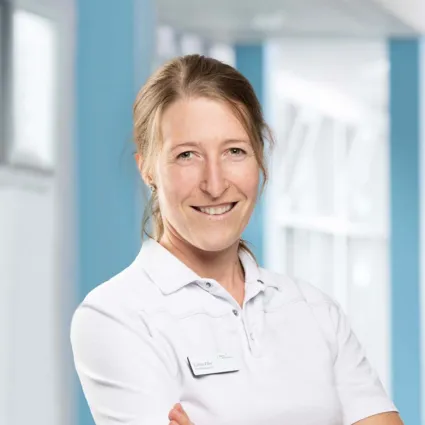 Lächelnde Frau mit langen, blonden Haaren, trägt ein weißes Polohemd und hat eine Namensschilder auf der linken Brustseite. Der Hintergrund ist hell und zeigt eine moderne, lichtdurchflutete Umgebung.
