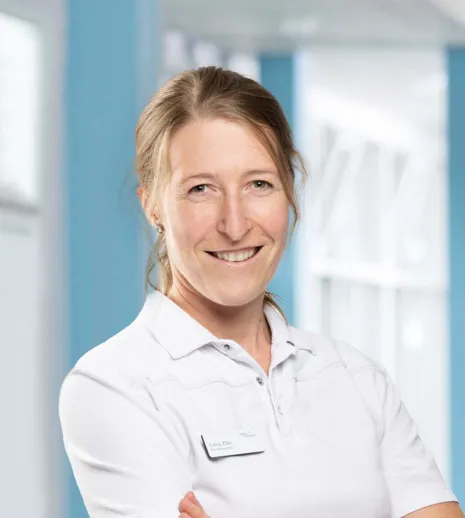 Lächelnde Frau mit langen, blonden Haaren, trägt ein weißes Polohemd und hat eine Namensschilder auf der linken Brustseite. Der Hintergrund ist hell und zeigt eine moderne, lichtdurchflutete Umgebung.