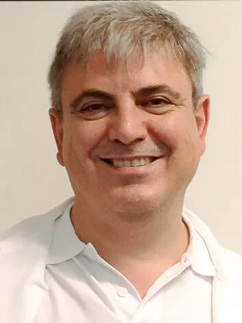 Porträt eines lächelnden Mannes mit kurzen grauen Haaren und weißem Hemd vor hellem Hintergrund.