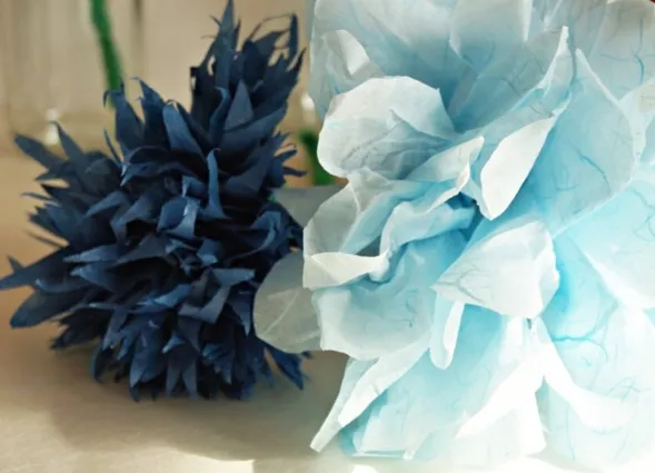 Zwei dekorative Papierblumen in Blau und Hellblau, eine mit spitzen Blütenblättern und die andere mit sanften, geschichteten Formen. Im Hintergrund sind unscharfe Glasbehälter sichtbar.