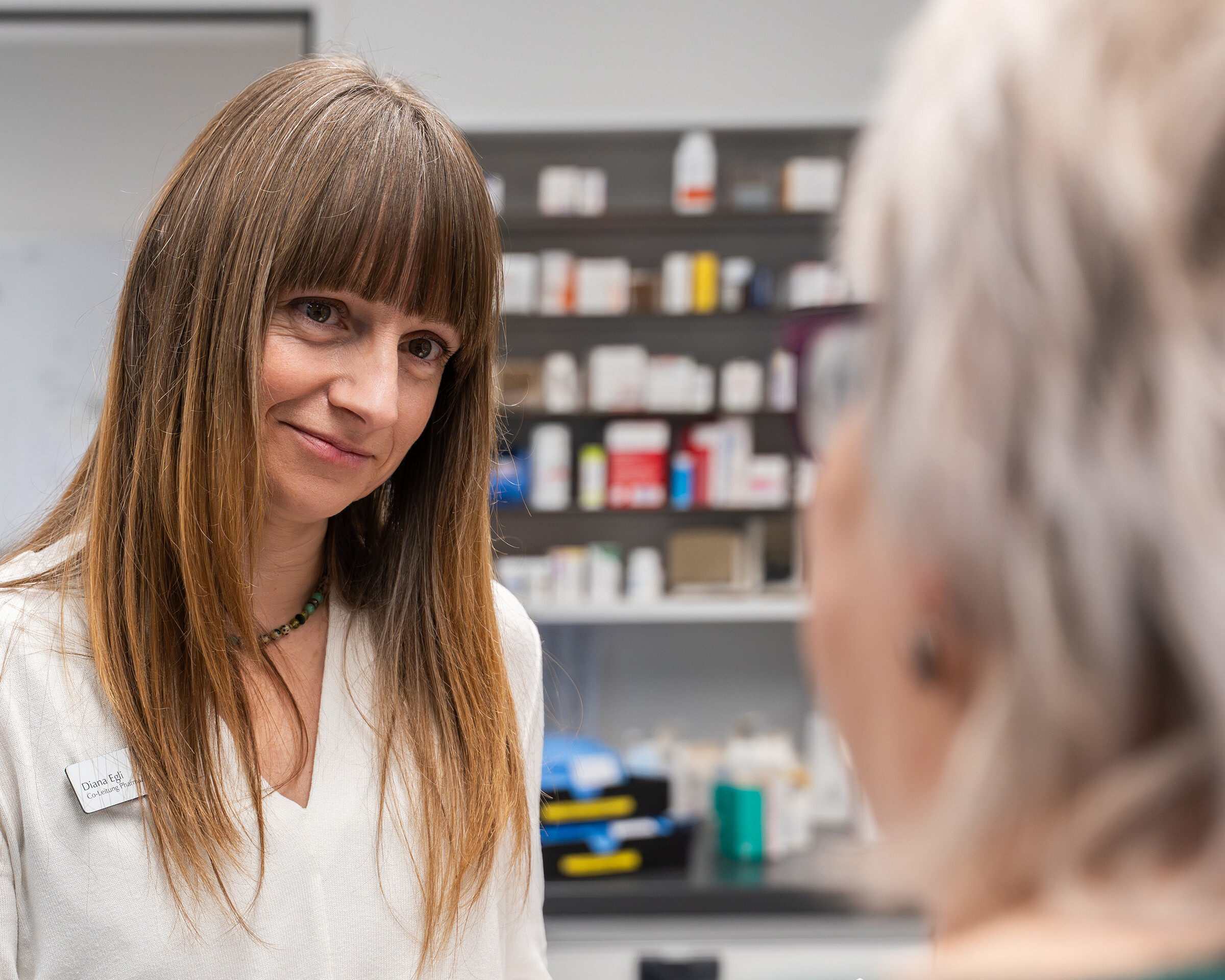 Freundliche Apothekerin im weißen Kittel im Gespräch mit einer älteren Kundin; Regale mit Medikamenten im Hintergrund.
