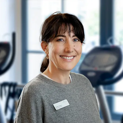 Lächelnde Frau mit braunem Haar in Zopfform, grauem Pullover mit Namensschild, im Hintergrund ein Fitnessstudio mit Geräten.