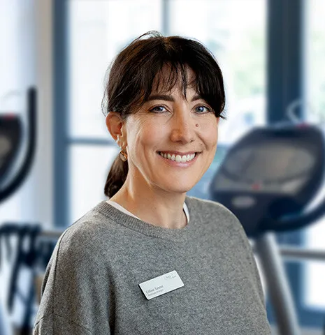 Lächelnde Frau mit braunem Haar in Zopfform, grauem Pullover mit Namensschild, im Hintergrund ein Fitnessstudio mit Geräten.