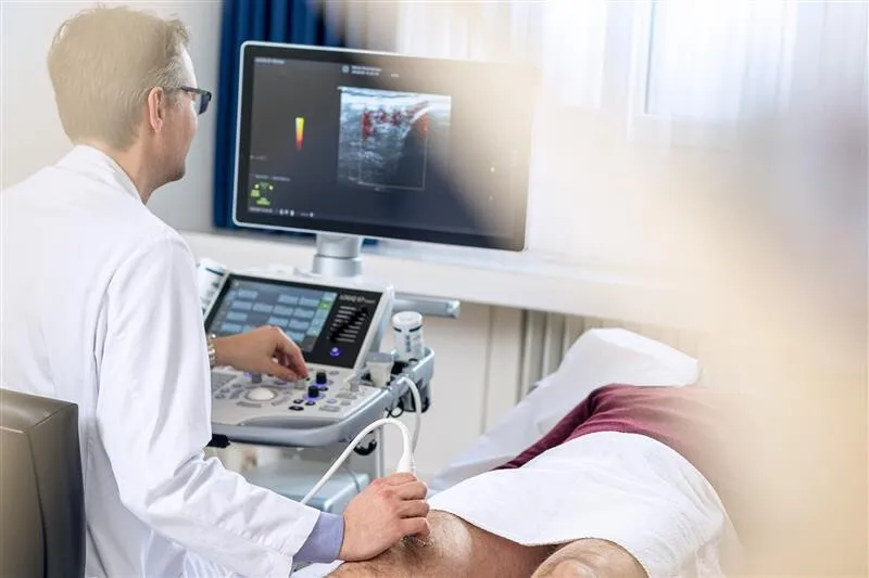 Arzt in weißem Kittel führt eine Ultraschalluntersuchung am Bein eines Patienten durch, während ein Monitor mit Ultraschallbild sichtbar ist.