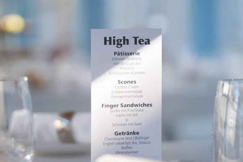 Gedeckter Tisch mit Gläsern, Tassen, Besteck und Menükarte für High Tea in einem hellen Café.