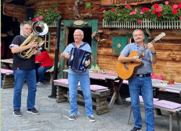 Drei Musiker auf einem Dorfplatz vor einer Holzfassade: links Tuba, in der Mitte Akkordeon, rechts E-Gitarre; im Hintergrund Blumenampeln und Bänke, dekorierte Tische.