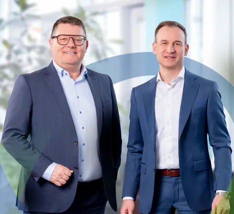Zwei Männer in Anzügen mit Hemden stehen nebeneinander und lächeln vor einem Büro-Hintergrund; im Hintergrund ist ein großes, halbtransparentes Logo mit grünen und blauen Linien erkennbar.
