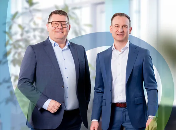 Zwei Männer in Anzügen mit Hemden stehen nebeneinander und lächeln vor einem Büro-Hintergrund; im Hintergrund ist ein großes, halbtransparentes Logo mit grünen und blauen Linien erkennbar.