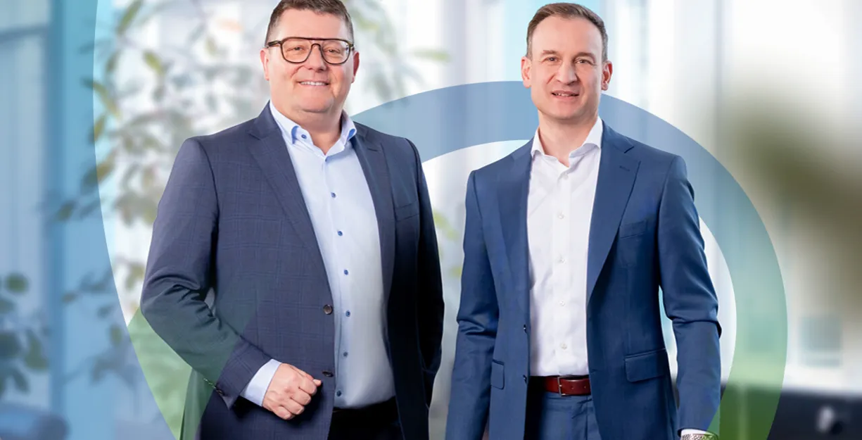 Zwei Männer in Anzügen mit Hemden stehen nebeneinander und lächeln vor einem Büro-Hintergrund; im Hintergrund ist ein großes, halbtransparentes Logo mit grünen und blauen Linien erkennbar.