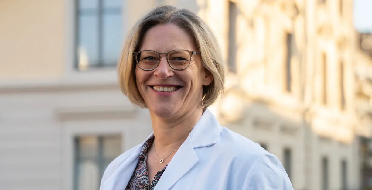 Lächelnde Frau mit kurzen, blonden Haaren und Brille, trägt einen weißen Laborkittel. Im Hintergrund sind helle Gebäude und Fenster zu sehen, die eine städtische Umgebung andeuten.