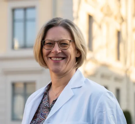 Lächelnde Frau mit kurzen, blonden Haaren und Brille, trägt einen weißen Laborkittel. Im Hintergrund sind helle Gebäude und Fenster zu sehen, die eine städtische Umgebung andeuten.