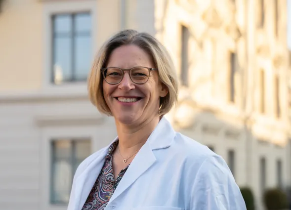 Lächelnde Frau mit kurzen, blonden Haaren und Brille, trägt einen weißen Laborkittel. Im Hintergrund sind helle Gebäude und Fenster zu sehen, die eine städtische Umgebung andeuten.