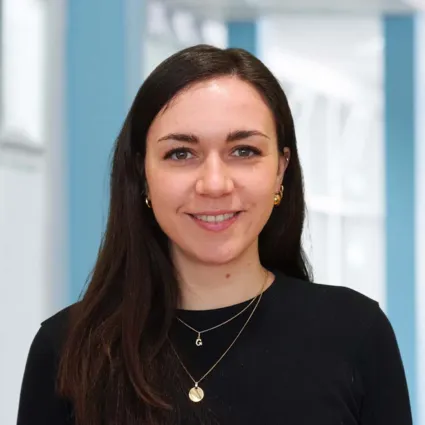 Frau mit langen, dunklen Haaren und einem freundlichen Lächeln, trägt ein schwarzes Oberteil und goldene Ohrringe, vor einem hellblauen Hintergrund.