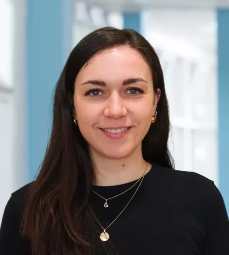 Frau mit langen, dunklen Haaren und einem freundlichen Lächeln, trägt ein schwarzes Oberteil und goldene Ohrringe, vor einem hellblauen Hintergrund.