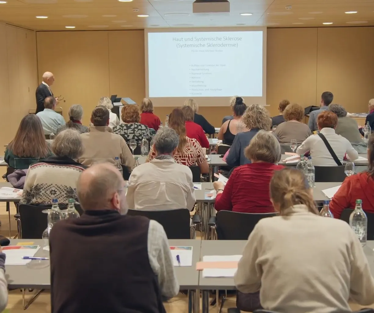 Teilnehmer eines Seminars sitzen in einem Raum, während ein Referent an einer Leinwand mit dem Thema 