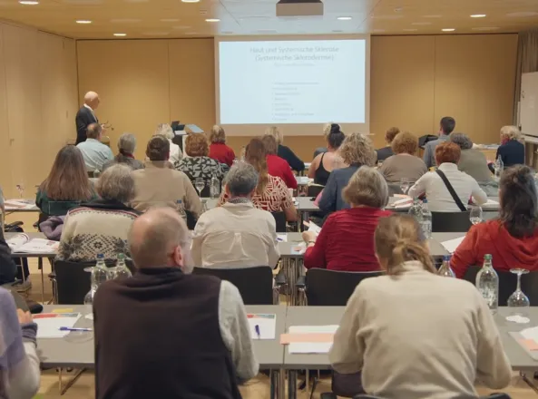 Teilnehmer eines Seminars sitzen in einem Raum, während ein Referent an einer Leinwand mit dem Thema 
