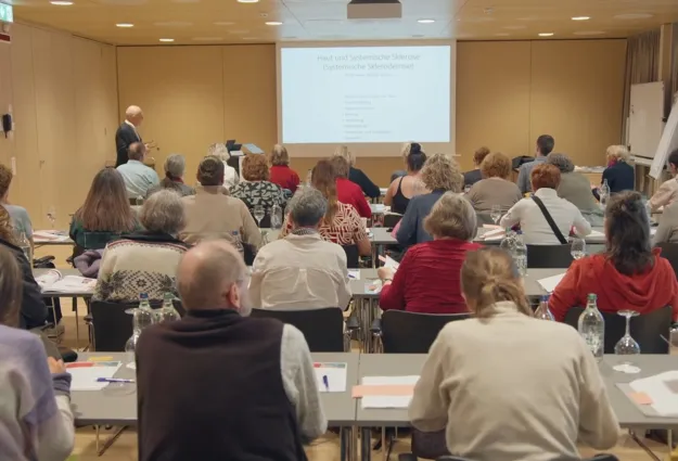 Teilnehmer eines Seminars sitzen in einem Raum, während ein Referent an einer Leinwand mit dem Thema 