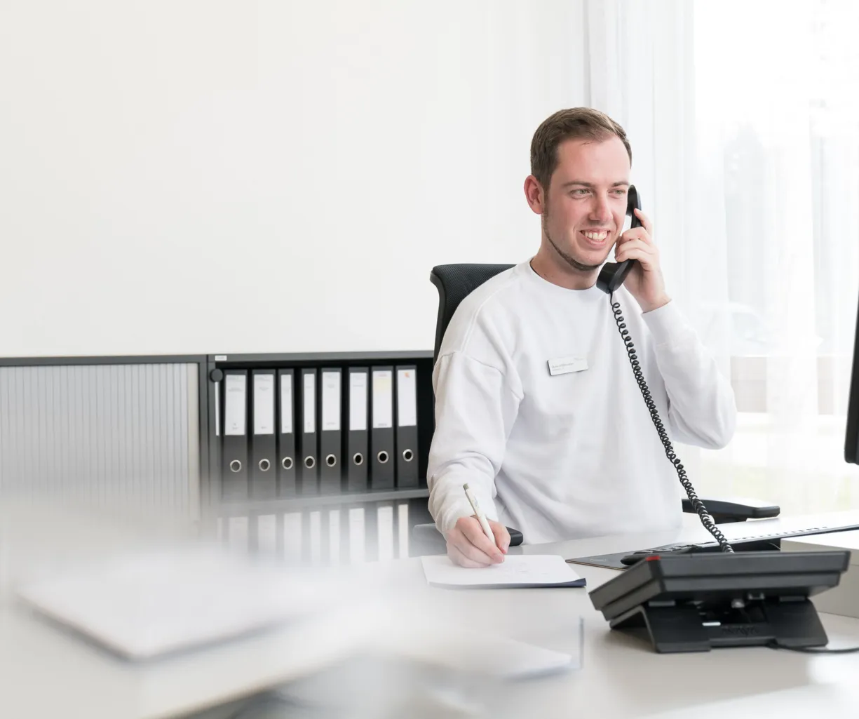 Mann am Schreibtisch mit Telefon, lächelnd, trägt ein weißes Sweatshirt, notiert etwas auf einem Blatt Papier. Im Hintergrund sind Aktenordner und ein Computer sichtbar.