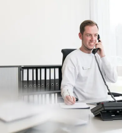 Mann am Schreibtisch mit Telefon, lächelnd, trägt ein weißes Sweatshirt, notiert etwas auf einem Blatt Papier. Im Hintergrund sind Aktenordner und ein Computer sichtbar.