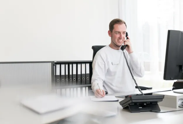 Mann am Schreibtisch mit Telefon, lächelnd, trägt ein weißes Sweatshirt, notiert etwas auf einem Blatt Papier. Im Hintergrund sind Aktenordner und ein Computer sichtbar.