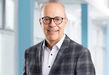 Mann mit Brille und grauem karierten Blazer, lächelt vor einem hellen, modernen Hintergrund mit blauen Akzenten.