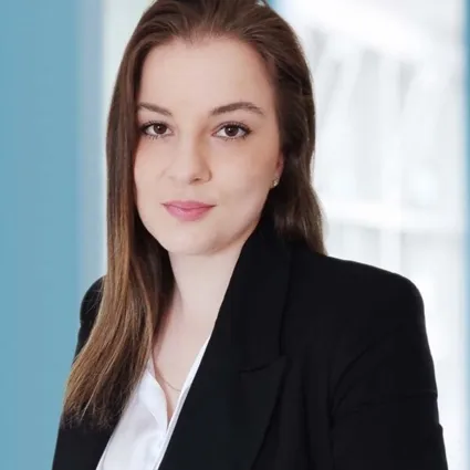 Portrait einer Frau im Business-Look mit dunkelbraunem Haar, schwarzer Blazerjacke und weißem Hemd, vor unscharfem blau-hellen Hintergrund.