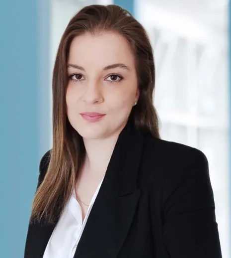 Portrait einer Frau im Business-Look mit dunkelbraunem Haar, schwarzer Blazerjacke und weißem Hemd, vor unscharfem blau-hellen Hintergrund.