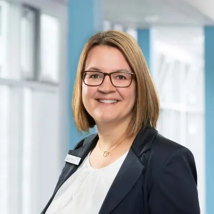 Frau mit schulterlangen, braunen Haaren und Brille, trägt einen schwarzen Blazer über einem weißen Oberteil, lächelt freundlich vor einem hellblauen Hintergrund mit Fenster.