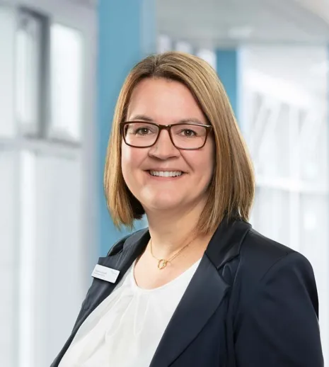 Frau mit schulterlangen, braunen Haaren und Brille, trägt einen schwarzen Blazer über einem weißen Oberteil, lächelt freundlich vor einem hellblauen Hintergrund mit Fenster.