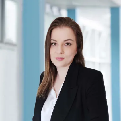 Frau mit langen, braunen Haaren in einem schwarzen Blazer und weißem Oberteil, stehend vor einem hellen, modernen Hintergrund mit blauen Akzenten.