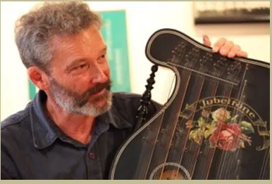 Bärtiger Mann betrachtet eine große, kunstvoll verzierte Zither mit Blumenmotiv und hält sie hoch.