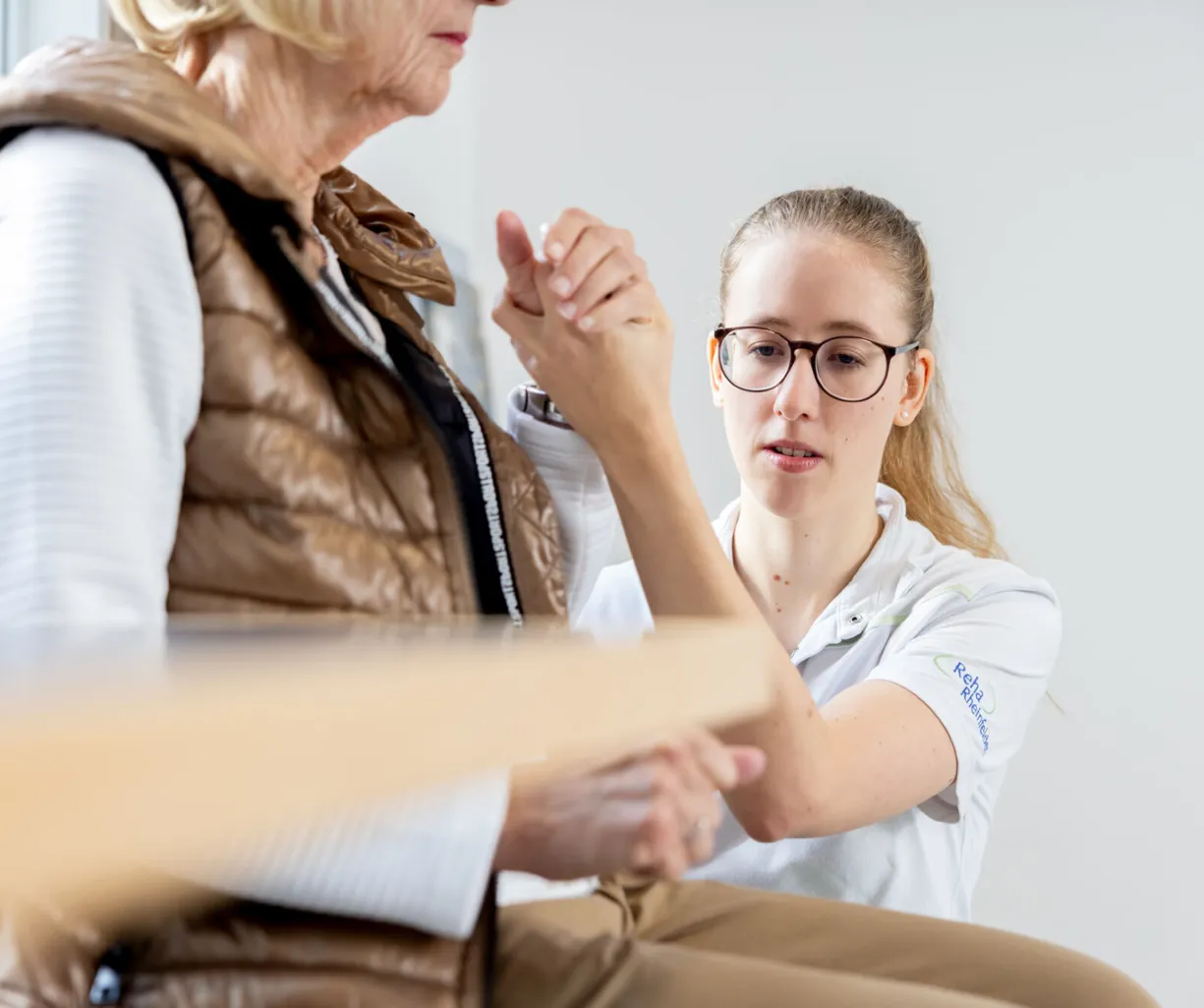 Eine junge Frau in einem weißen Poloshirt führt eine Handuntersuchung an einer älteren Frau durch, die eine braune Weste trägt. Beide sitzen in einem hellen Raum mit minimalistischem Hintergrund.