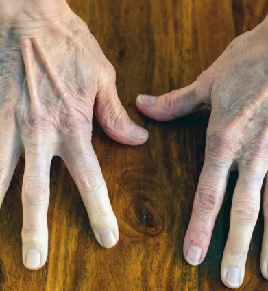 Zwei Hände mit sichtbaren Adern und Falten liegen auf einer Holzoberfläche, die Finger sind gespreizt. Die Nägel sind kurz und unlackiert.