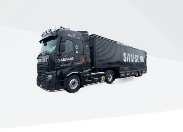 Schwarzer Lkw mit abgedecktem Ladecontainer, seitlich „SAMSUNG“, auf weißem Hintergrund mit schrägen Linien, dreiviertel Ansicht.