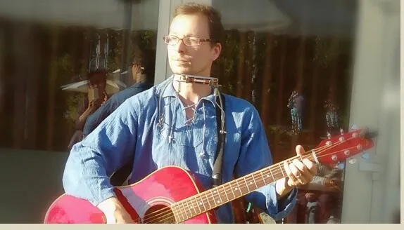 Mann mit Brille spielt eine rote Akustikgitarre im Licht einer Spiegelung, vor einem Fenster mit Vorhängen.