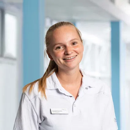 Lächelnde Mitarbeiterin in weißer Arbeitskleidung mit Namensschild vor hellem, modernem Hintergrund.