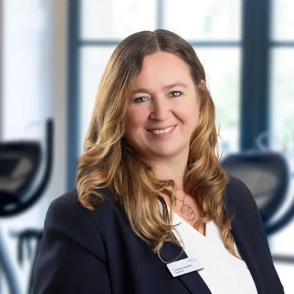 Porträt einer lächelnden Frau im Business-Blazer vor Fitnessgeräten in einem hellen Raum mit großen Fenstern.