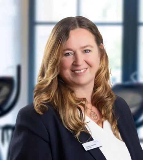 Porträt einer lächelnden Frau im Business-Blazer vor Fitnessgeräten in einem hellen Raum mit großen Fenstern.