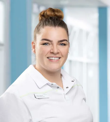 Lächelnde Frau mit hochgestecktem Haar, trägt ein weißes Poloshirt mit grünen Akzenten. Im Hintergrund sind helle, moderne Räumlichkeiten sichtbar.