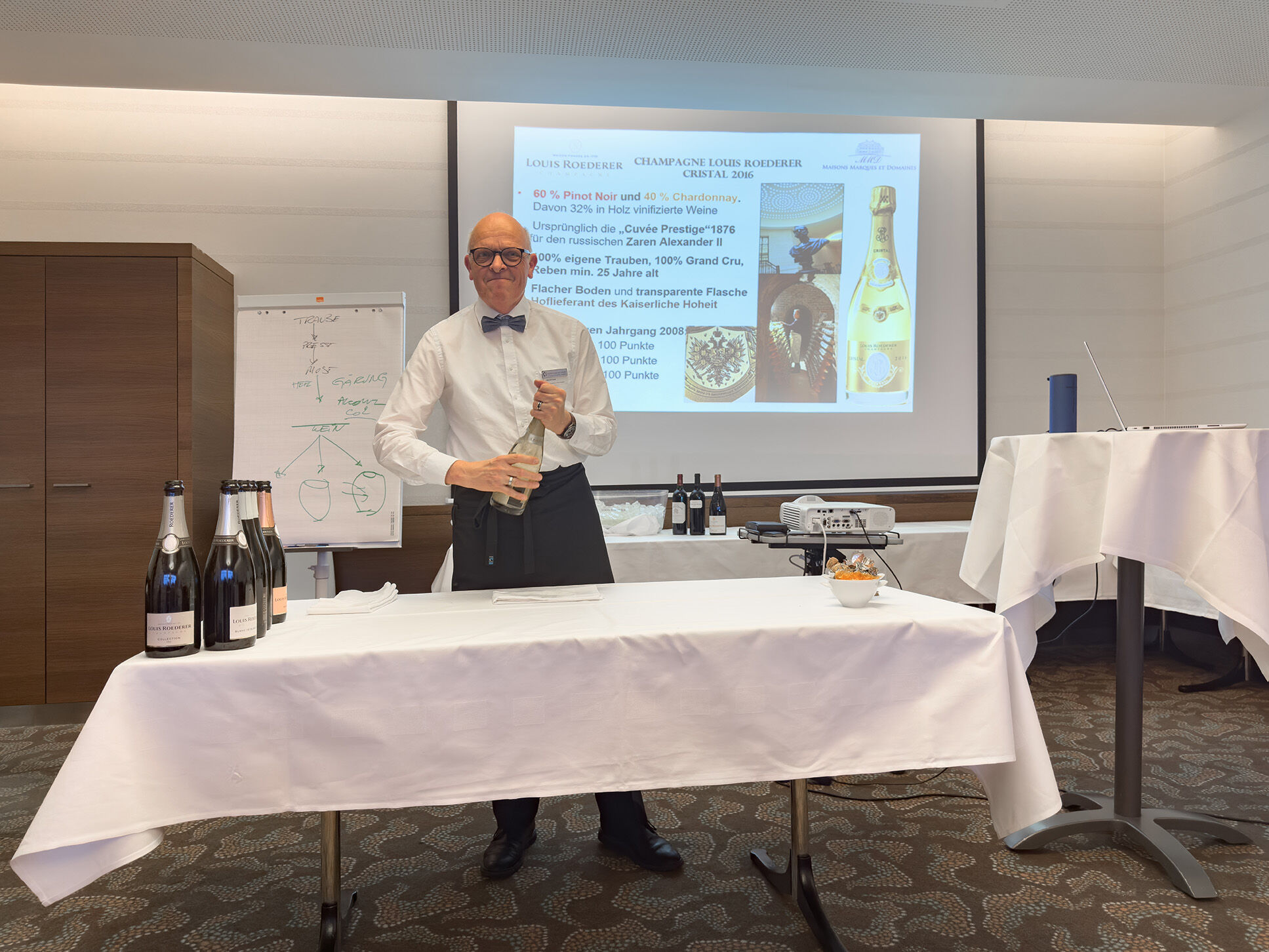 Ein Mann in einem weißen Hemd mit Fliege öffnet eine Flasche Champagner. Auf dem Tisch stehen mehrere Flaschen Champagner und ein Notizblock. Im Hintergrund ist eine Präsentation über Louis Roederer und Champagner zu sehen. Der Raum ist hell und modern...