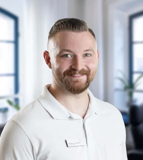Porträt eines lächelnden Mannes mit kurzem Haar und Bart im weißen Poloshirt, vor hellen Fenstern in einem Büro.