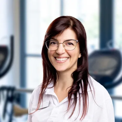 Porträt einer Frau mit braunen Haaren und Brille in weißem Hemd vor unscharfem Büro-Hintergrund.