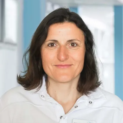 Frau mit langen, dunklen Haaren und einem freundlichen Gesichtsausdruck, trägt ein weißes Oberteil. Im Hintergrund sind unscharfe, helle Wände zu sehen.