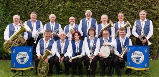 Musikkapelle im Freien: mehrere Musiker im blauen Anzug mit Instrumenten wie Trompeten, Posaunen, Saxofon und Schlagzeug, im Hintergrund Hecke; zwei große blaue Schilder mit Emblem.